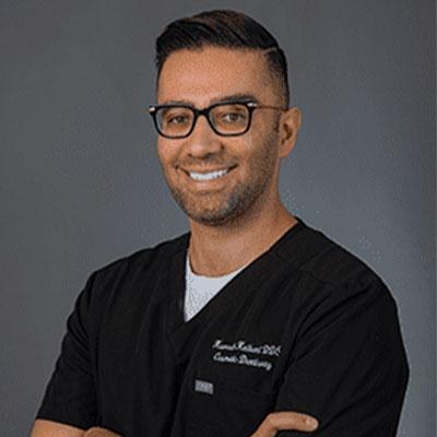 Portrait of a smiling man with glasses wearing dark scrubs against a gray background